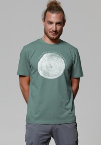 Grünes Baumwoll-T-Shirt mit einem weißen runden Grafikdesign, das einem Baumring ähnelt, mit Text um das Muster herum. Kurze Ärmel und Rundhalsausschnitt.