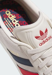 White suede sneaker featuring red and blue stripes, gold text, and a padded tongue. The interior displays a colorful brand logo pattern.