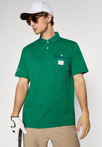 Polo verde din bumbac. Include un buzunar cu etichetă, trei nasturi și mâneci scurte. Asortat cu pantaloni bej și o pălărie albă.