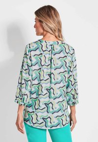 Bluse mit einem bunten geometrischen Muster in Grün, Blau und Schwarz. Sie hat Dreiviertelärmel und einen runden Ausschnitt mit einem kleinen Knopfverschluss.