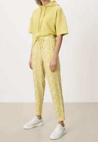 Gul hættebluse med korte ærmer, parret med lette, blomster mønstrede gule bukser. Modellen bærer hvide sneakers.