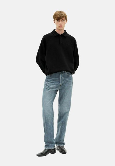 Juodas megztinis su plokščiu apykakle ir trimis mygtukais, derinamas su šviesiai mėlynomis tiesiomis kelnėmis ir juodais batais.