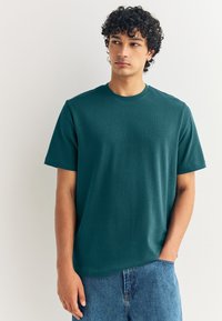 Dunkelgrünes Baumwoll-T-Shirt mit Rundhalsausschnitt und kurzen Ärmeln, das eine leichte Struktur und eine entspannte Passform aufweist. Kombiniert mit blauen Jeans.