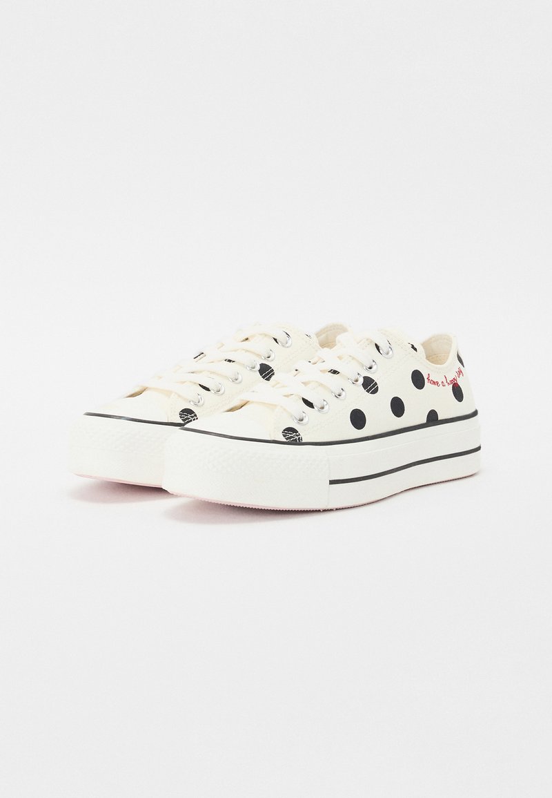 Zapatillas de lona blanca con lunares negros, acentos de suela negra y rayas negras. Cuentan con punta redonda y cordones planos.