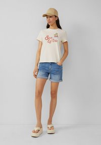 Crème T-shirt met korte mouwen met de grafische tekst "Sea La Vie", gecombineerd met blauwe denim shorts, een beige pet en platform sandalen.