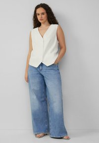 Weißes, geripptes ärmelloses Hemd mit V-Ausschnitt und Knopfdetails, kombiniert mit hellblauen, weit geschnittenen Jeans und silbernen Sandalen.