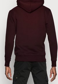 Sudadera burdeos con capucha ajustable, puños y dobladillo acanalados. Textura suave, diseño ajustado y un color sólido sin logotipos visibles.