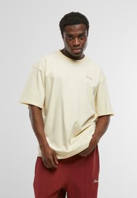 Korte mouwen, crème T-shirt gemaakt van katoen. Heeft een ronde halslijn en een subtiele geborduurde logo op de linkerkant van de borst. G gedragen met rode broek.