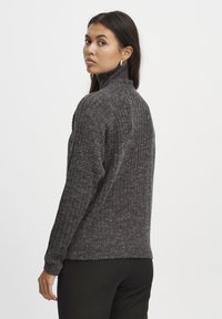 Siv turtleneck pulover z ribano teksturo, prilegajočimi se rokavi in sproščenim krojem, v kombinaciji z črnimi hlačami.