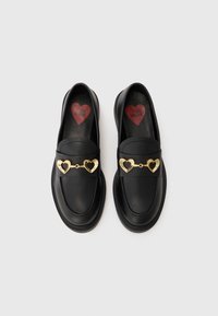 Μαύρα loafers με λείο δερμάτινο φινίρισμα, με χρυσή αλυσίδα σε σχήμα καρδιάς. Στο εσωτερικό, ένα κόκκινο λογότυπο καρδιάς στη σόλα.