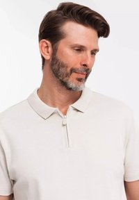 Světlobéžové polo tričko s jemnou texturou, krátkými rukávy a detaily s zipovým límcem. Látka vypadá prodyšně a uvolněně.