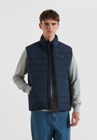 Marineblaues Steppweste mit hohem Kragen, horizontalen Steppmustern und Seitentaschen, über einem grauen Sweatshirt getragen. Goldfarbener Logoakzent.
