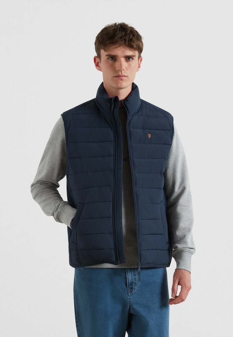 Marineblaues Steppweste mit hohem Kragen, horizontalen Steppmustern und Seitentaschen, über einem grauen Sweatshirt getragen. Goldfarbener Logoakzent.