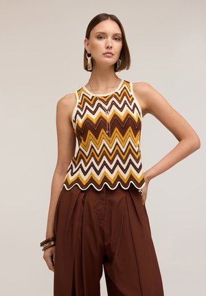 Femme portant un top en maille sans manches à motif zigzag marron, jaune et blanc avec un ourlet festonné, associé à un pantalon marron taille haute et des bijoux en or.