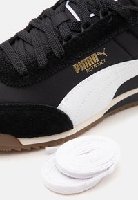 Μαύρο αθλητικό παπούτσι Puma Retrojet με λευκή ρίγα και χρυσό λογότυπο, μαύρα κορδόνια και επιπλέον λευκά σπειροειδή κορδόνια σε λευκό φόντο.
