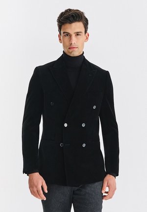 Mężczyzna w czarnym dwurzędowym żakiecie i czarnym swetrze z golfem, stojący na jednolitym białym tle.