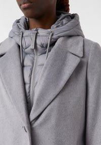 Gros plan sur une personne portant un manteau en laine gris clair superposé à une veste grise matelassée à fermeture éclair avec capuche à cordon.