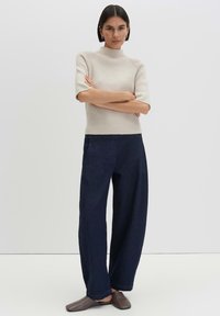 Woman with dark hair stands with arms crossed wearing a beige short-sleeve sweater, dark wide-leg pants, and brown slip-on shoes.