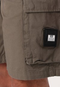 Shorts cargo vert olive en coton, dotés d'une poche à patch avec une étiquette "WEEKEND OFFENDER" en noir et blanc et des détails de couture visibles.