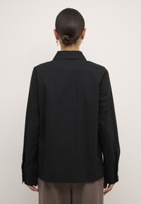 Camicia nera a maniche lunghe con colletto a punta, realizzata in tessuto morbido. Presenta polsini con bottoni e una vestibilità rilassata, vista dal retro.
