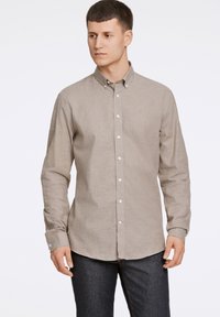Camicia button-up marrone chiaro con maniche lunghe, colletto strutturato, bottoni bianchi e un tessuto morbido e testurizzato. Indossata con jeans scuri.