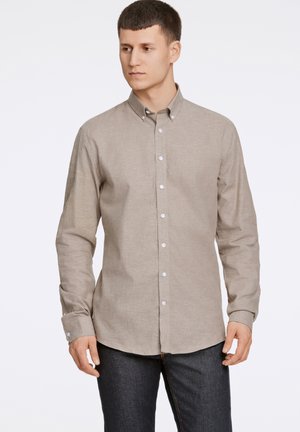 Camisa de botón color marrón claro con mangas largas, cuello estructurado, botones blancos y una tela suave y texturizada. Llevada con jeans oscuros.