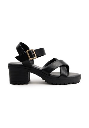 Sandalias de tacón negro con suela gruesa, con correas cruzadas y una hebilla ajustable. Fabricadas en cuero suave con un tacón texturizado.