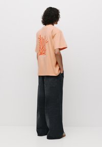 Tricou oversize piersică cu un grafic coral și text pe spate, asortat cu blugi negri cu picioare wide-leg și sandale cu vârf deschis.