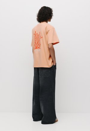 Fersken oversized t-shirt med en koral grafisk tryk og tekst på ryggen, parret med vide sorte jeans og åbne sandaler.