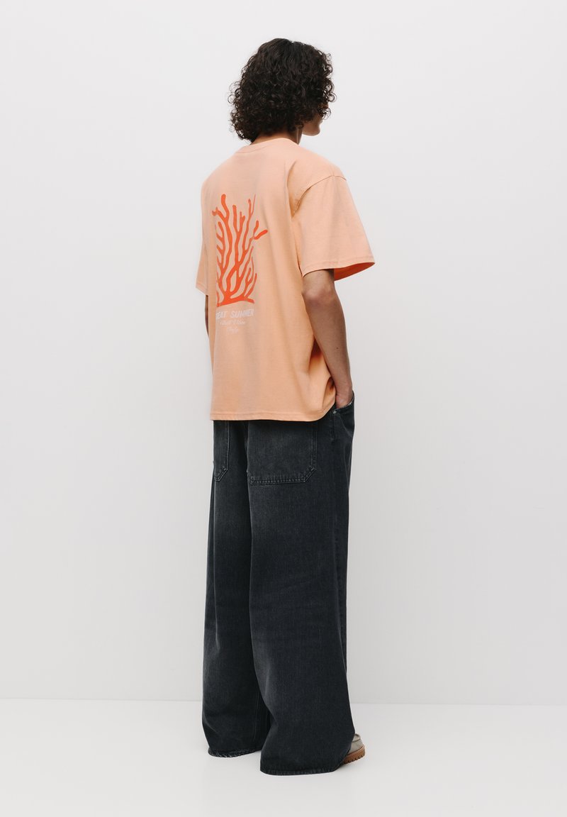 Tricou oversize piersică cu un grafic coral și text pe spate, asortat cu blugi negri cu picioare wide-leg și sandale cu vârf deschis.