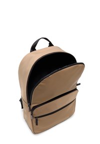 Sac à dos beige avec une texture lisse et mate, accents de fermeture éclair noirs, poches à double fermeture éclair et une poignée supérieure pour le transport.