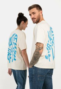 Krämfarbendes T-Shirt mit kurzen Ärmeln, das auf der Rückseite großen blauen Text zeigt. Kombiniert mit blauen Jeans, die einen lässigen Schnitt und zeitgemäßes Design präsentieren.