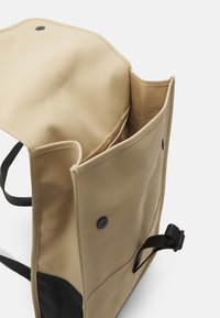 Beige väska med svart botten, med en lockstängning, knappknäppning och fickor inuti. Tillverkad av hållbart material med en slät textur.
