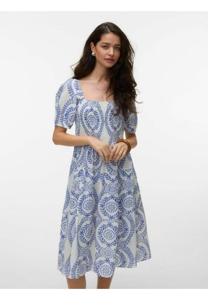 Kvinde med langt mørkt hår iført en hvid midi-kjole med blå cirkulære blomsterprint og puffede korte ærmer.