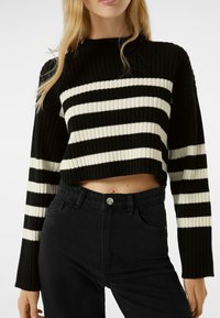 Czarnobiały sweter w paski, krótki, o prążkowanej fakturze, z długimi rękawami i okrągłym dekoltem, zestawiony z czarnymi, wysokim stanem dżinsami.