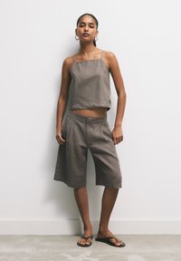 Hnědý lněný cropped top s tenkými ramínky a odpovídajícími šortkami ke kolenům, se sklady a volným střihem. Na sobě má černé žabky.