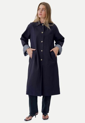 Femme portant un long manteau bleu marine avec doublure à carreaux aux poignets, pantalon noir et sandales noires à talons ouverts, debout les mains dans les poches du manteau.