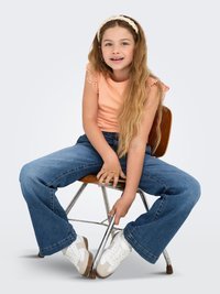 Hellbeige Kurzarmtop mit Spitzenakzenten, blaue Weitbundjeans, weiße Sneakers mit braunen Sohlen, sitzend auf einem Holzstuhl.