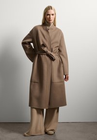 Lungo cappotto in morbida lana beige con cintura, colletto alto, due tasche frontali e maniche ampie. Indossato sopra pantaloni beige a gamba larga coordinati.