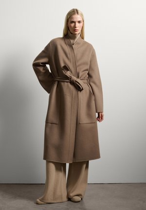 Lang, met riem, jas van zacht beige wol met een hoge kraag, twee voorzakken en brede mouwen. Gekocht over bijpassende beige broek met wijdere pijpen.