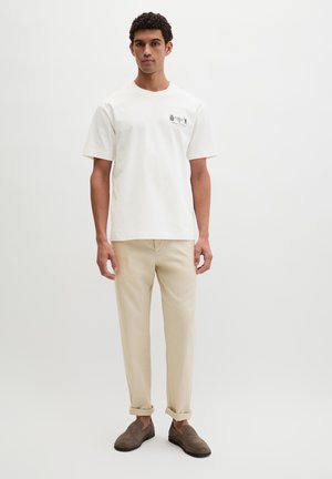 Homme portant un t-shirt blanc à motif graphique, un pantalon beige retroussé et des chaussures marron sans lacets, debout devant un fond clair uni.