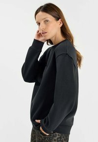 Svart sweatshirt med avslappnad passform, ribbade muddar och mjuk textur. Modellen bär den med mönstrade byxor, vilket visar en sidoprofil.