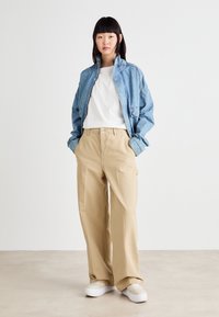 La veste en denim présente une coupe décontractée avec une couleur bleu clair, associée à un pantalon beige à jambes larges et des baskets blanches. Un simple t-shirt blanc en dessous.