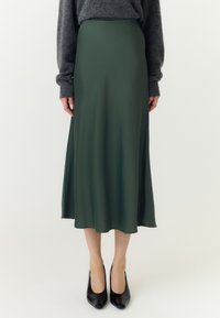 Kvinde iført en mørkegrøn midi-nederdel, grå langærmet sweater og sorte høje hæle, der står mod en ensfarvet lys baggrund.