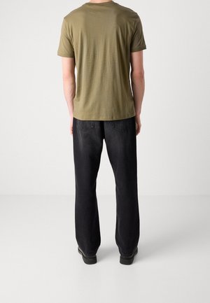 Mann trägt olivgrünes T-Shirt und weit geschnittene schwarze Jeans, steht mit dem Rücken zur Kamera vor einem schlichten weißen Hintergrund.