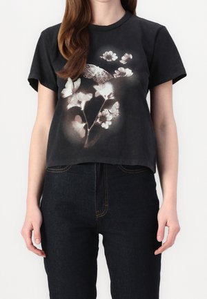 Frau trägt ein schwarzes T-Shirt mit kurzen Ärmeln mit einem verblassten Schmetterlings- und Blumenprint und dunkelblaue Jeans.