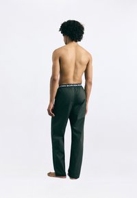 Calças verde-escuras com um corte descontraído e cintura elástica, apresentando um rótulo preto em contraste. Tecido suave, design até ao tornozelo.