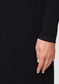 Zwart gebreid kledingstuk met een ribbeltextuur en een aansluitende pasvorm, met een close-up van een hand die op de stof rust.