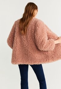 Giacca in pelliccia sintetica di un rosa tenue, caratterizzata da una forma ampia, maniche larghe e una superficie strutturata. Indossata su jeans blu scuro.