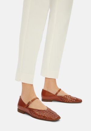 Zapatos planos de cuero marrón con diseño tejido en la punta. Presentan una correa en el tobillo, un tacón bajo y una textura suave. Combinados con pantalones blancos ajustados.